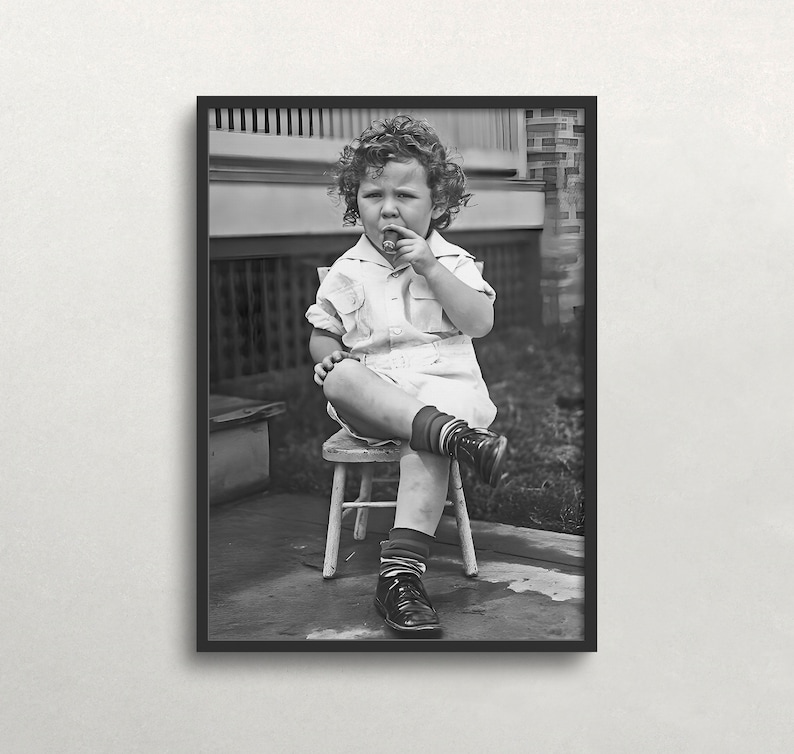Peut inclure: Une photo en noir et blanc d'un jeune enfant assis sur une petite chaise en bois, portant une chemise blanche et un short, et tenant une sucette dans la bouche.