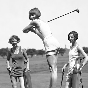May include: Three women on a golf course, two holding golf clubs and one holding a golf bag. The woman in the center is wearing a white tank top and white shorts. The woman on the left is wearing a red tank top and the woman on the right is wearing a white tank top and white shorts.