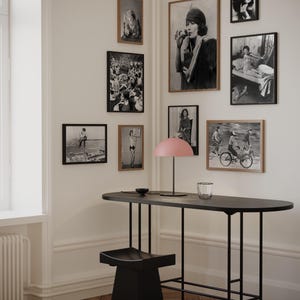 May include: A black metal table with a black stool and a pink lamp. The table has a long, oval top and a simple, modern design. The wall behind the table is decorated with black and white framed photographs.
