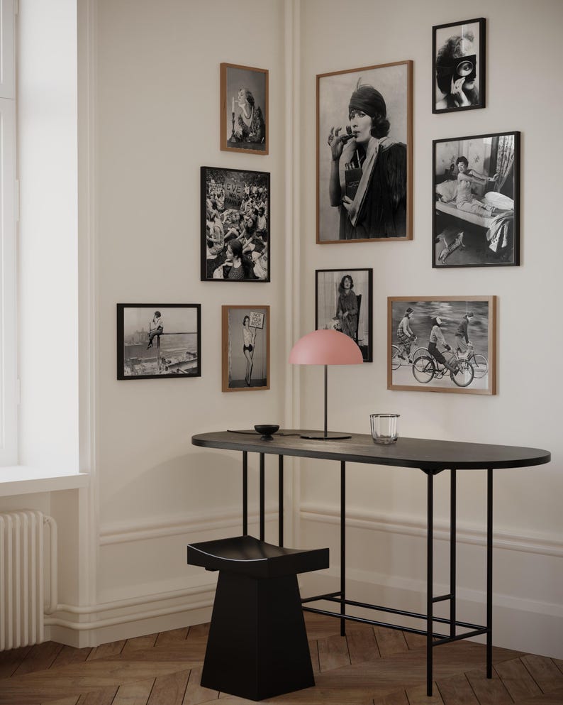 May include: A black metal table with a black stool and a pink lamp. The table has a long, oval top and a simple, modern design. The wall behind the table is decorated with black and white framed photographs.