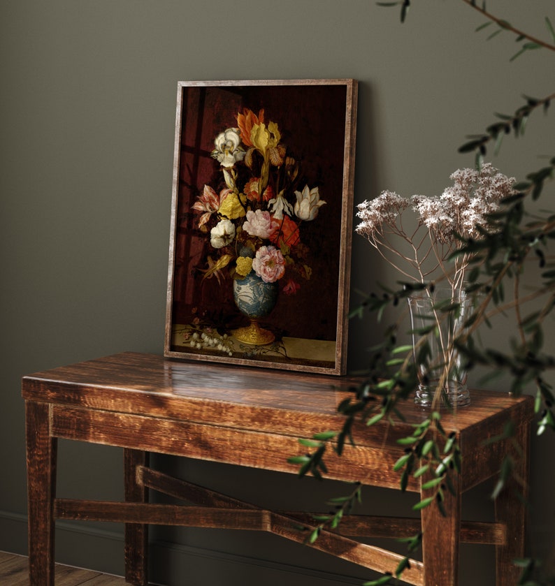 May include: A framed floral still life painting featuring a vase filled with colorful flowers. The vase is blue and white, set against a dark red background. The painting is displayed on a rustic wooden table.