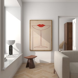 May include: A framed wall art print featuring a minimalist red line drawing of a martini glass with a pair of red lips resting on the rim, set against a light beige background, displayed on a white wall above a small brown wooden stool.