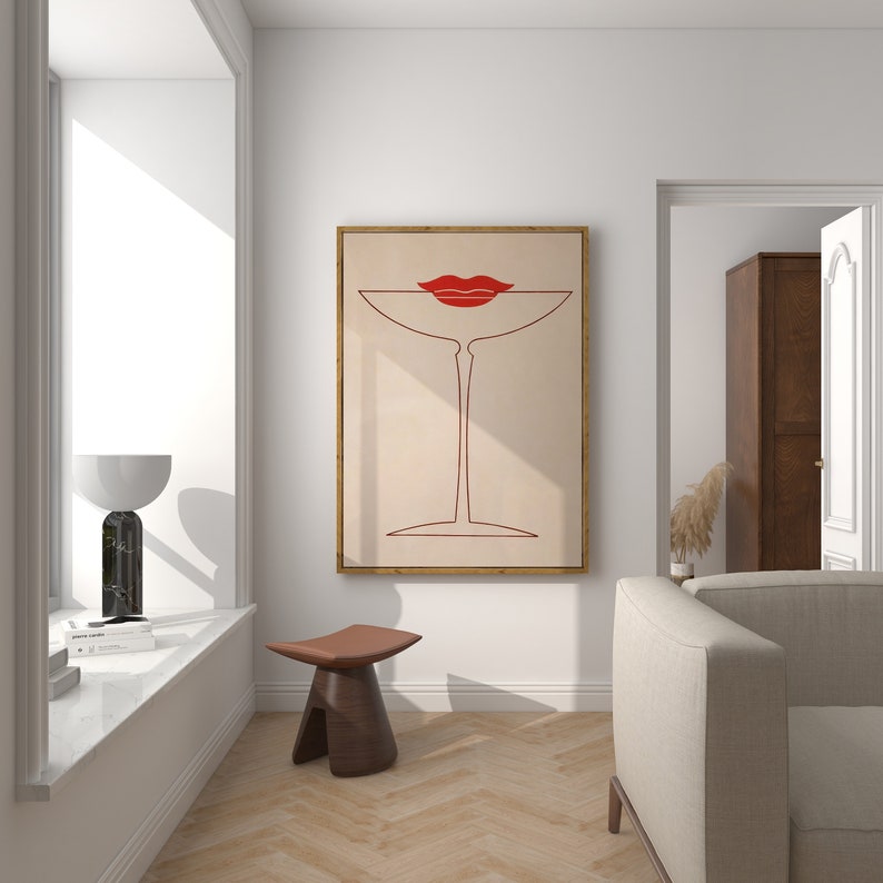 May include: A framed wall art print featuring a minimalist red line drawing of a martini glass with a pair of red lips resting on the rim, set against a light beige background, displayed on a white wall above a small brown wooden stool.