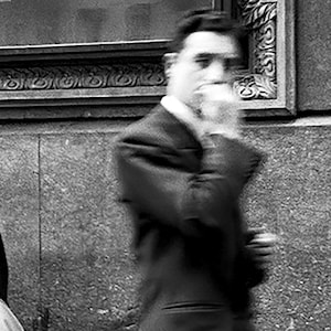 Pu&ograve; includere: Fotografia in bianco e nero di una persona in abito, in movimento sfocato. La persona &egrave; in piedi vicino a un edificio con dettagli architettonici. L'immagine &egrave; granulosa, suggerendo una fotografia pi&ugrave; vecchia.