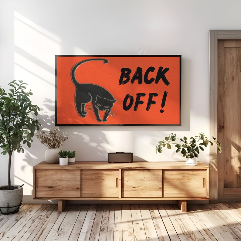 May include: A black cat illustration on an orange background with the text "BACK OFF!" displayed on a framed wall art. The artwork is above a wooden cabinet with three sliding doors. A potted plant is on the left.
