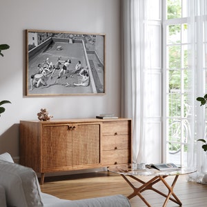 Puede incluir: Una impresi&oacute;n en blanco y negro enmarcada representa a un grupo de personas en una azotea. La obra de arte cuelga sobre un mueble de madera con una puerta tejida. Una mesa de centro con tapa de cristal y un sof&aacute; blanco est&aacute;n en primer plano. Cortinas blancas transparentes enmarcan una ventana.