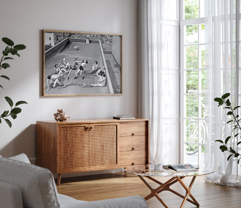 Puede incluir: Una impresi&oacute;n en blanco y negro enmarcada representa a un grupo de personas en una azotea. La obra de arte cuelga sobre un mueble de madera con una puerta tejida. Una mesa de centro con tapa de cristal y un sof&aacute; blanco est&aacute;n en primer plano. Cortinas blancas transparentes enmarcan una ventana.