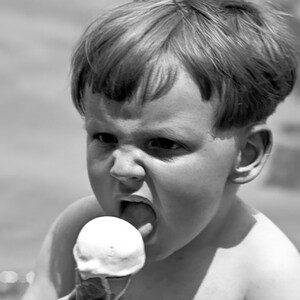 May include: A young boy with short, light-colored hair is eating a white ice cream cone. He is looking at the camera with a serious expression on his face.