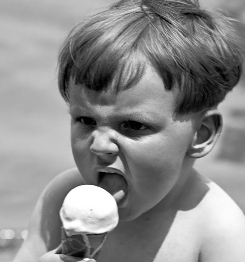 May include: A young boy with short, light-colored hair is eating a white ice cream cone. He is looking at the camera with a serious expression on his face.