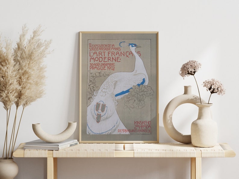 Op de afbeelding: Een ingelijste vintage art nouveau poster met een witte pauw met blauwe accenten. De poster, met Franse tekst, adverteert een kunsttentoonstelling in Praag in 1902. Het wordt getoond op een houten bank met decoratieve vazen en gedroogde planten.