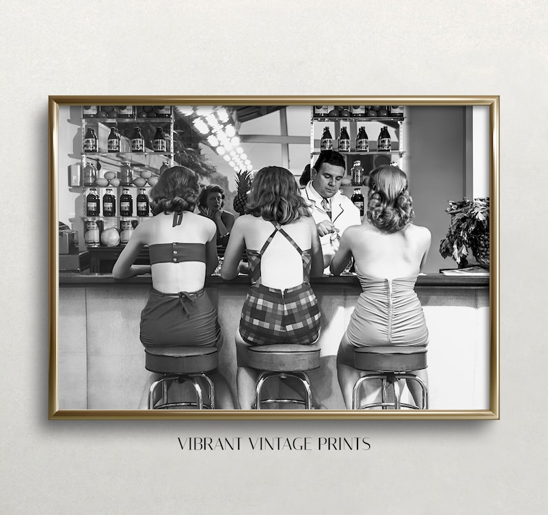 Op de afbeelding: Zwart-witfoto van drie vrouwen die aan een bar zitten in badpakken. De vrouwen kijken naar de camera en een man staat achter de bar. De bar heeft een rij flessen erachter.