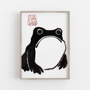 May include: A framed print featuring a minimalist black and white illustration of a frog. The frog has large eyes and a wide mouth, with a red stamp in the upper left corner. The artwork is set against a white background.
