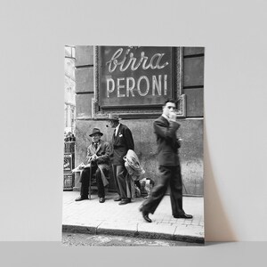 Pu&ograve; includere: Fotografia in bianco e nero di una scena di strada con tre uomini. Un'insegna al neon recita "birra PERONI". Un uomo fuma la pipa, un altro &egrave; seduto su una panchina e un terzo passa camminando.