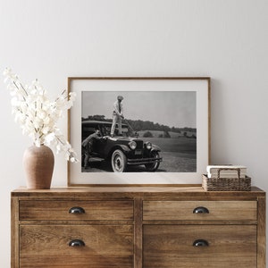 Op de afbeelding: Zwart-witfoto van twee mannen die naast een vintage auto staan. De man op de auto houdt een golfclub vast. De auto heeft een kentekenplaat met de tekst "61-517".