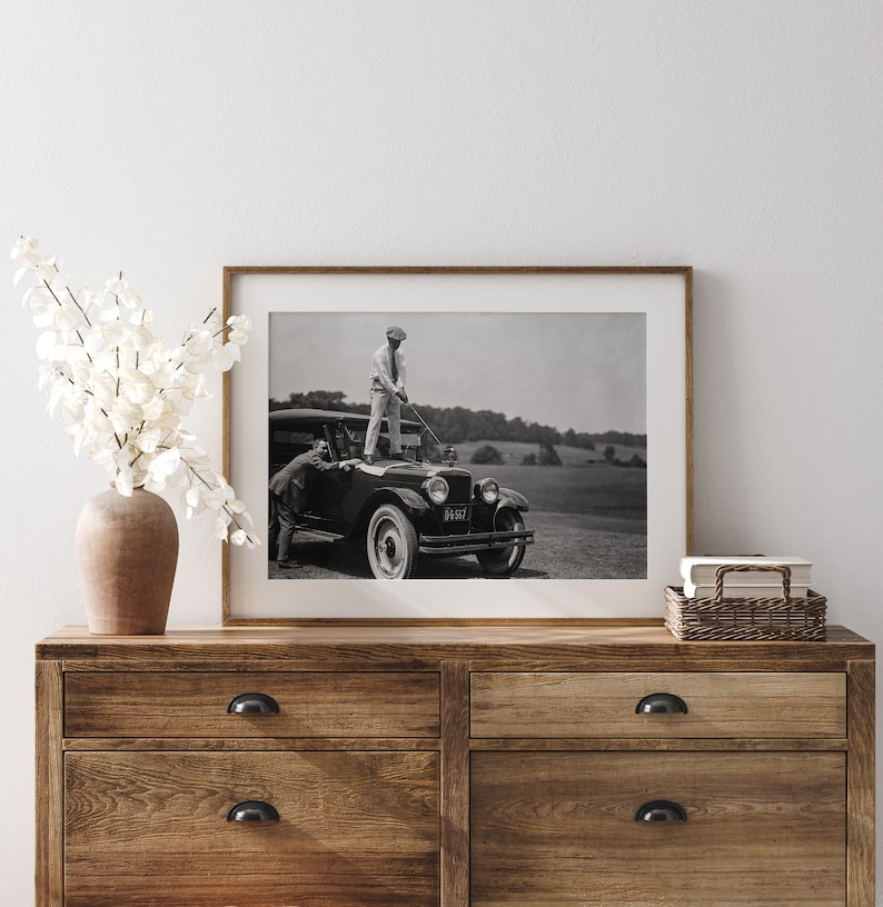 Op de afbeelding: Zwart-witfoto van twee mannen die naast een vintage auto staan. De man op de auto houdt een golfclub vast. De auto heeft een kentekenplaat met de tekst "61-517".