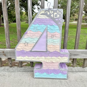 May include: A large, colorful number four piñata, perfect for a birthday celebration. The piñata is decorated with layers of pastel-colored crepe paper in shades of purple, blue, yellow, and pink. It has a silver crown and star accents.
