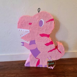 May include: Pink dinosaur piñata with purple and pink details. The piñata has a white paper mouth with teeth and a purple eye.
