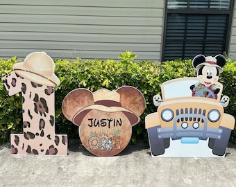 Può includere: Tre sagome di cartone per una festa di primo compleanno. La prima sagoma è un grande numero uno con un design a stampa leopardata. La seconda sagoma è una testa di Topolino con un cappello da safari e il testo "Justin Wild One". La terza sagoma è una jeep gialla con Topolino al volante.
