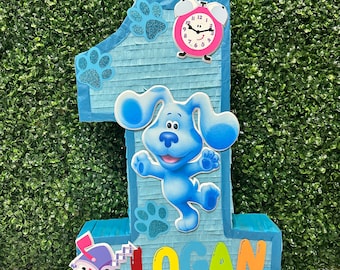 May include: Blue's Clues themed piñata shaped like the number one. The piñata is blue with a white border and features a blue dog character, a pink clock, and the name "LOGAN" in colorful letters.