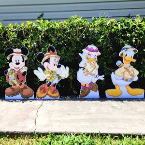 May include: Four cardboard stand-up figures of Disney characters dressed in safari outfits. Mickey Mouse, Minnie Mouse, Donald Duck, and Daisy Duck are wearing khaki shorts, shirts, and hats. The characters are standing on a sidewalk in front of a green bush.