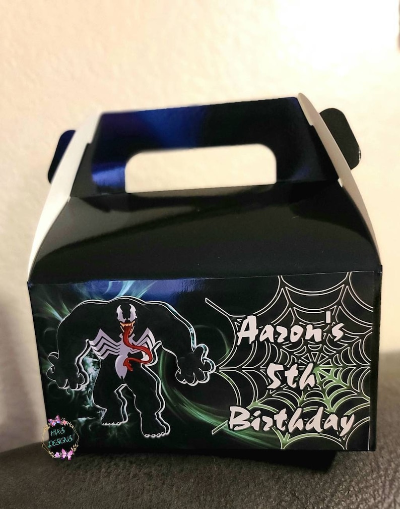 May include: Black and green favor box with a cartoon image of Venom, a Marvel Comics character. The box has a white handle and says "Aaaron's 5th Birthday" on the front.