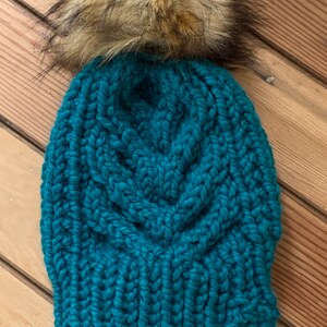 May include: Teal knitted beanie with a large brown and white faux fur pom pom on top.