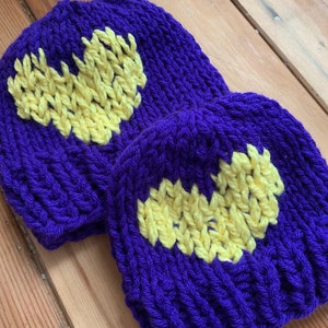 May include: Two knitted beanies, one large and one small, in a vibrant purple color. Each beanie features a yellow heart design on the front. The hats are made of a thick, textured yarn.