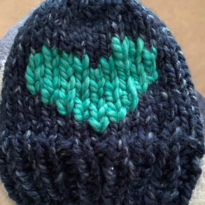 May include: A dark blue knit hat with a teal heart design. The hat is made of a thick, textured yarn with flecks of white throughout. The heart is prominently displayed in the center of the hat.