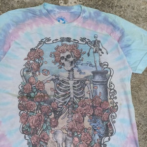 May include: Tie-dye t-shirt featuring a skeleton design adorned with roses, set within an ornate frame. The shirt's tie-dye pattern includes pastel shades. The collar displays the Liquid Blue logo.