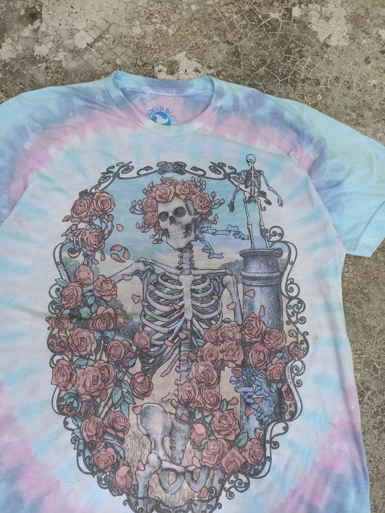 May include: Tie-dye t-shirt featuring a skeleton design adorned with roses, set within an ornate frame. The shirt's tie-dye pattern includes pastel shades. The collar displays the Liquid Blue logo.