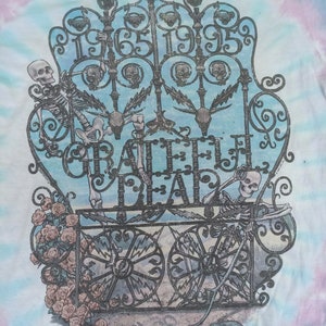 May include: Tie-dye t-shirt with a detailed graphic of a wrought iron gate, the words "Grateful Dead", and the years "1965-1995". The design includes skeletons, roses, and a turtle. The background has a blue, pink, and white tie-dye pattern.