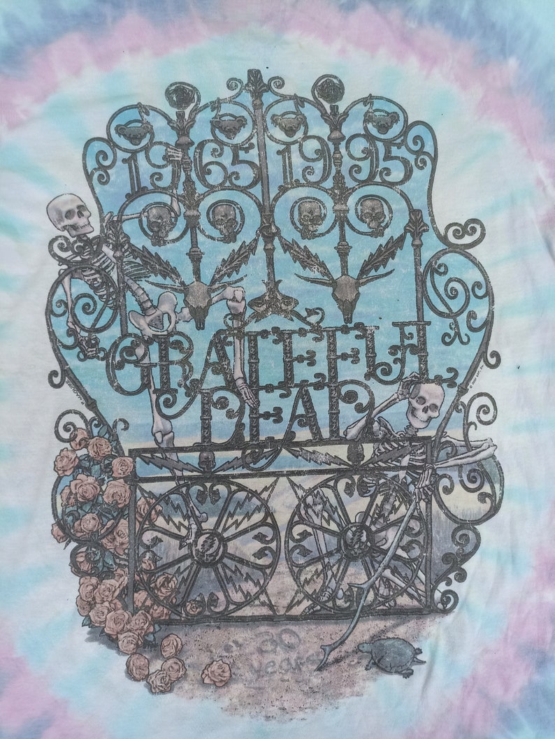 May include: Tie-dye t-shirt with a detailed graphic of a wrought iron gate, the words "Grateful Dead", and the years "1965-1995". The design includes skeletons, roses, and a turtle. The background has a blue, pink, and white tie-dye pattern.
