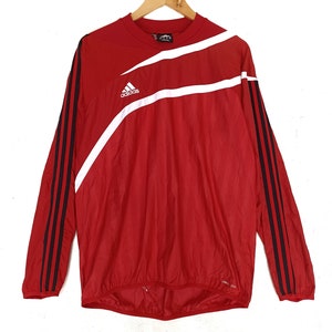 Puede incluir: Camiseta deportiva Adidas roja de manga larga con rayas diagonales blancas y rayas negras en las mangas. El logo de Adidas está en el pecho. La camiseta está colgada en una percha de madera.