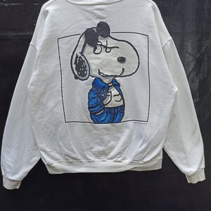 Peut inclure: Sweat-shirt blanc avec une illustration en noir et blanc de Snoopy portant une veste en jean bleu et un jean. L'illustration est encadrée dans un rectangle noir.