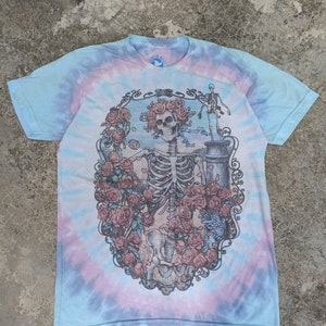 May include: A tie-dye t-shirt with a blue, pink, and white spiral pattern. The front features a detailed graphic of a skeleton surrounded by roses. The shirt has short sleeves and a crew neck, with the "Liquid Blue" logo visible.