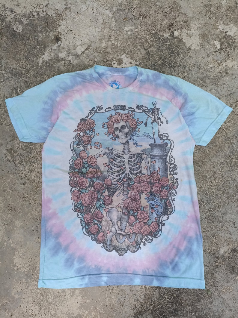 May include: A tie-dye t-shirt with a blue, pink, and white spiral pattern. The front features a detailed graphic of a skeleton surrounded by roses. The shirt has short sleeves and a crew neck, with the "Liquid Blue" logo visible.