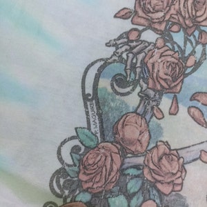 May include: Close-up of a light fabric with a floral design. The design features several pink roses and a skeletal hand. The background has a watercolour effect with shades of blue and green. The name "P. Maguire" is printed on the design.