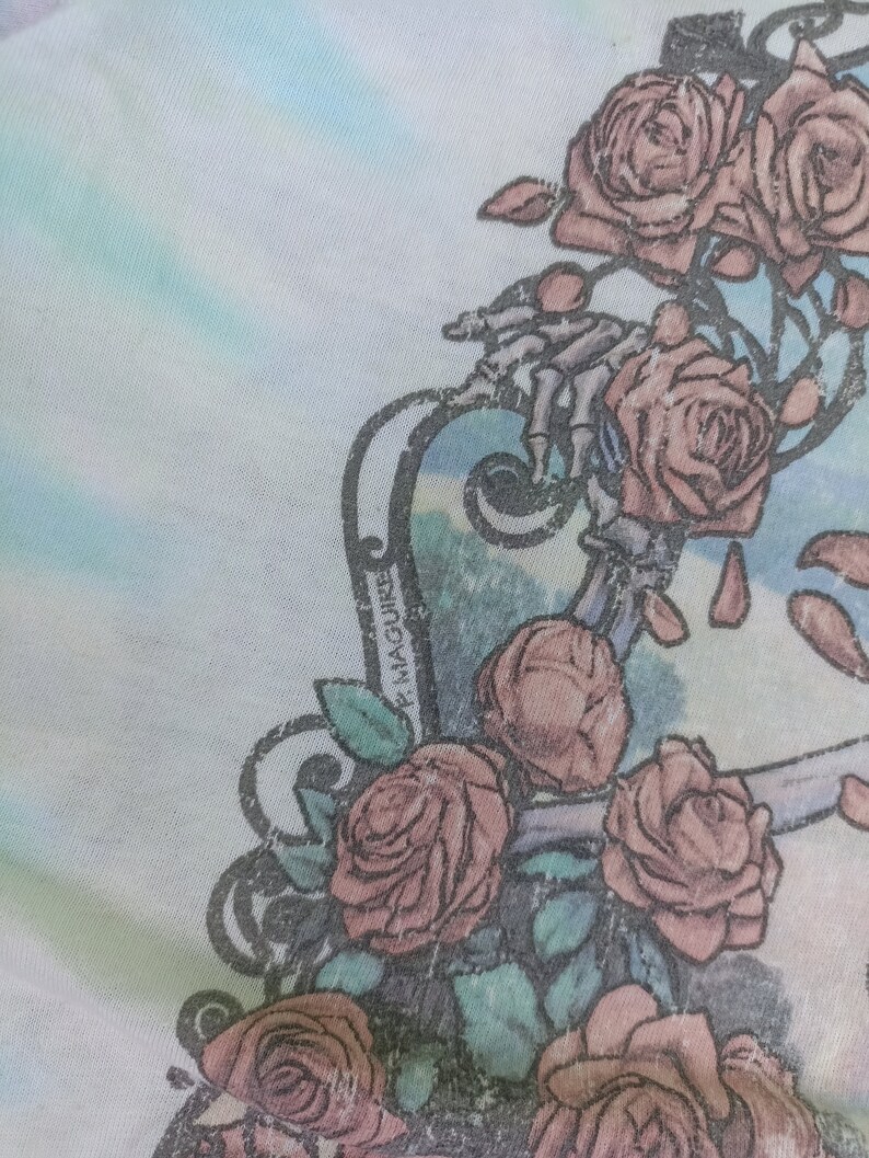 May include: Close-up of a light fabric with a floral design. The design features several pink roses and a skeletal hand. The background has a watercolour effect with shades of blue and green. The name "P. Maguire" is printed on the design.