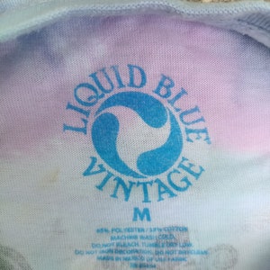 May include: Close-up of a tie-dye t-shirt with the "Liquid Blue Vintage" logo in blue. The shirt is medium size and made of a polyester and cotton blend. Washing instructions are printed below the logo.