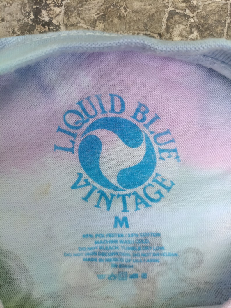 May include: Close-up of a tie-dye t-shirt with the "Liquid Blue Vintage" logo in blue. The shirt is medium size and made of a polyester and cotton blend. Washing instructions are printed below the logo.