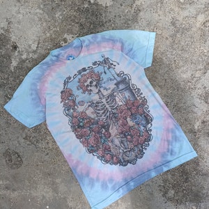 May include: A tie-dye t-shirt featuring a skeleton design encircled by roses. The shirt has a spiral pattern in blue, pink, and white. The graphic is centrally placed on the garment.