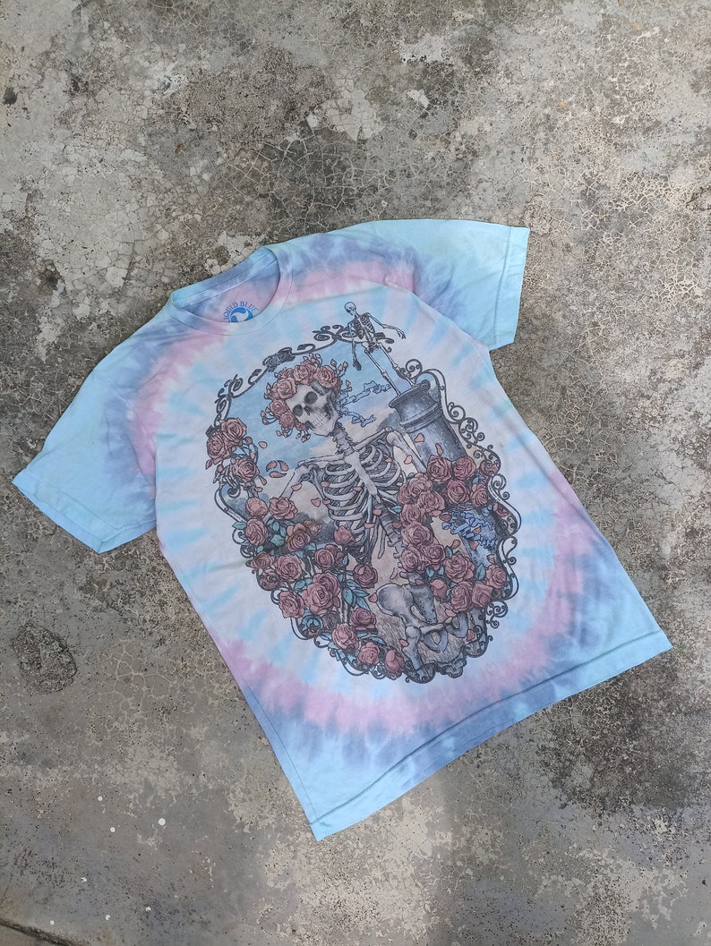 May include: A tie-dye t-shirt featuring a skeleton design encircled by roses. The shirt has a spiral pattern in blue, pink, and white. The graphic is centrally placed on the garment.