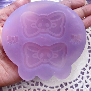 May include: A light purple silicone mold featuring a skull with a bow design. The mold has a rounded shape with a scalloped edge and includes two larger skull bow designs and two smaller bow designs.