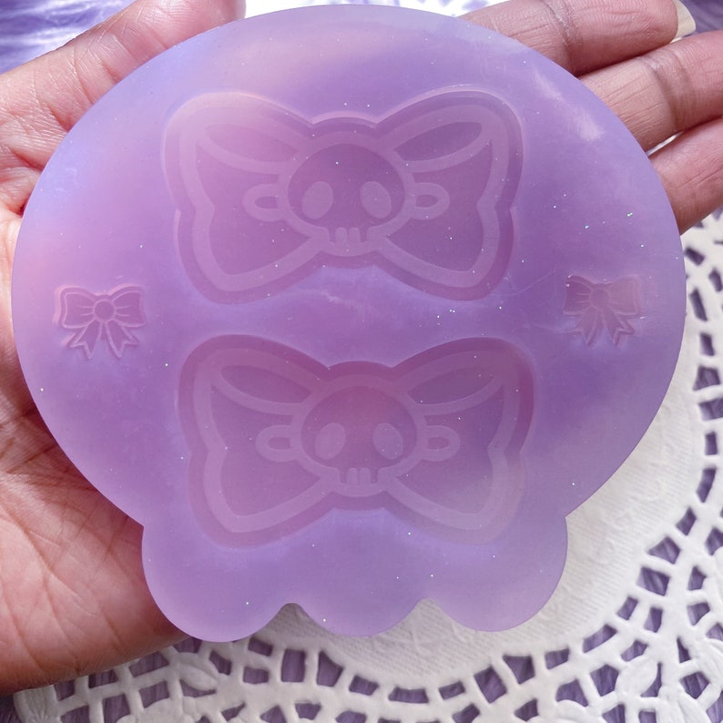 May include: A light purple silicone mold featuring a skull with a bow design. The mold has a rounded shape with a scalloped edge and includes two larger skull bow designs and two smaller bow designs.