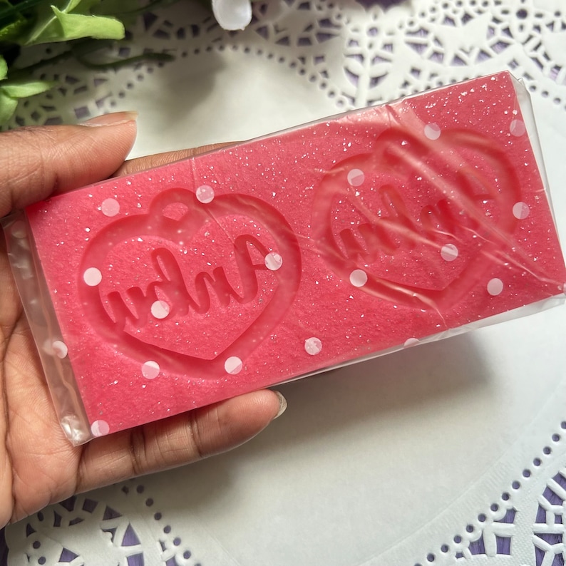 May include: Pink silicone mold with white polka dots. The mold has two heart shapes with the words "mama" and "dada" inside.
