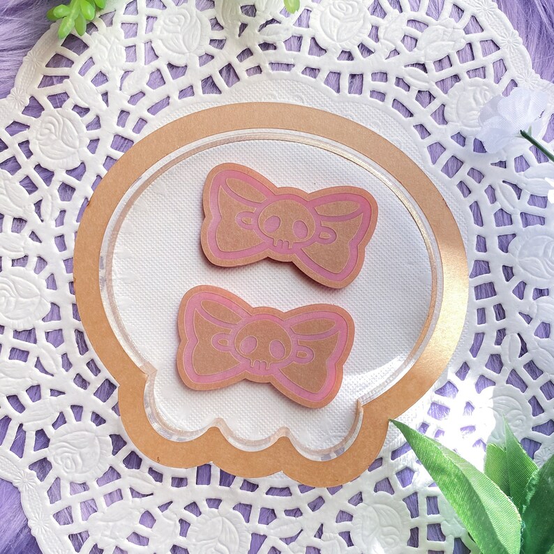 May include: Two stickers with a skull and bow design. The stickers are light brown with pink outlines and skull details. They are placed on a white doily with a brown border, with a purple background.
