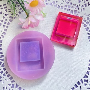 May include: Two silicone molds for resin casting. The larger mold is purple and has a square frame with a smaller square inside. The smaller mold is pink and has a square frame. Both molds are on a white lace doily.