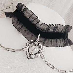 May include: A black choker necklace featuring a central velvet band with ruffled sheer fabric edges. A silver-toned metal pendant shaped like an ornate heart with a small cross inside hangs from the center, attached to a silver-toned chain.