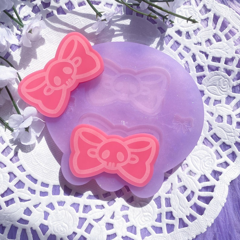 May include: A light purple silicone mold with a scalloped edge, featuring a bow design with a skull in the center. Two pink bow-shaped resin pieces with skull details are placed on top. The background includes white lace and purple fabric.