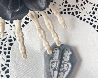 May include: A gray silicone mold in the shape of a coffin featuring two knife-shaped cavities. The mold is positioned beneath a skeletal hand prop and a black fabric flower with a single eye in the center, resting on a white paper doily.